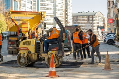 Construction workers in orange vests repair city road with vehicles during the day.